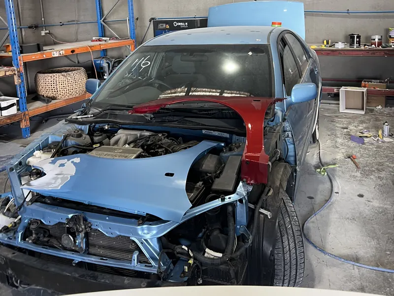 A light blue sedan in the workshop with its front end stripped for repair